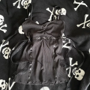 Svart handsydd korsett-topp med spets och snörning - Snygg svart korsett-topp med smala axelband och dekorativa rosetter. Toppen har ribbat tyg, spetsdetaljer och snörning i sidorna för extra edge. Perfekt för dig som gillar alternativ stil och vill sticka ut med coola detaljer. Plagget är handsytt, vilket innebär att det kan förekomma mindre skavanker tex lösa trådar! 🪡 