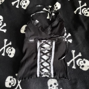 Svart handsydd korsett-topp med snörning och spets - Säljer en svart korsett-topp med ribbad nederdel, spetsdetaljer och snörning framtill. Toppen har halterneck med satinband att knyta i nacken och v-ringad spetskant. Snygg kontrast med vita detaljer längs snörningen. Perfekt för dig som gillar alternativ stil. Plagget är handsytt, vilket innebär att det kan förekomma mindre skavanker tex lösa trådar! 🪡 