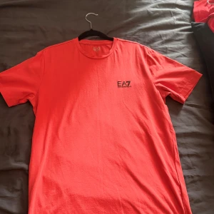 Röd EA7 Emporio Armani t-shirt - Snygg röd t-shirt från EA7 Emporio Armani med svart logga på bröstet. Klassisk rund halsringning och korta ärmar. Perfekt för dig som gillar stilrena och trendiga plagg med premiumkänsla. Skön passform och mjukt material.