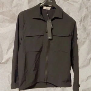 Svart overshirt från Stone Island - Svart overshirt från Stone Island med klassisk krage, två stora bröstfickor och dragkedja framtill. Jackan har den ikoniska Stone Island-loggan på ärmen och är tillverkad i ett lätt syntetmaterial. Perfekt för lager-på-lager och stilren streetwear. Jackan passar på folk mellan 175-185. För bilder när jag har på mig den skriv pm🤗 Äktenhetsbevis finns!