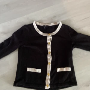 Svart kofta med vita detaljer från H&M - Stilren svart kofta från H&M med vita kanter och guldiga knappar framtill. Koftan har två dekorativa fickor och långärmad passform. Perfekt för dig som gillar klassisk och chic stil.