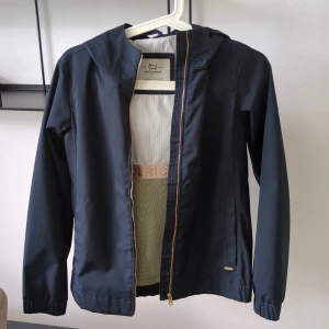 Mörkblå vindjacka från Woolrich - Snygg mörkblå vindjacka från Woolrich med huva och guldfärgad dragkedja.Knappt använd, ny pris 1800 kr.  Jackan är tillverkad i polyester och har ett stilrent, minimalistiskt uttryck. Insidan har diskreta ränder och meshdetaljer samt logoband. Perfekt för blåsiga dagar när du vill hålla stilen.