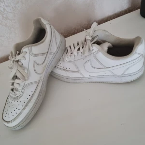 Nike Air Force 1 vita sneakers - Klassiska Nike Air Force 1 sneakers i helvitt skinn med perforerad tå, rund tå och platt sula. Snygg och tidlös modell med snörning och ikonisk swoosh på sidorna. Perfekta för dig som gillar streetstyle och vill ha en clean look.
