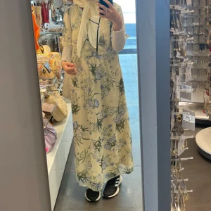 Blommig långklänning i ljusa toner - Superfin långklänning med v-ringning och korta ärmar. Klänningen är ljusbeige med blommigt mönster i grönt och lila. Den har en luftig passform och är perfekt för varma dagar. Materialet känns lätt och flowy, vilket gör den extra bekväm.