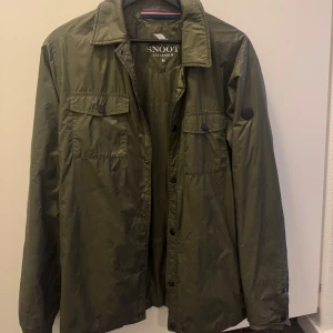 Grön SNOOT jacka - Snygg grön jacka från SNOOT i storlek M. Jackan har en klassisk krage och knappar framtill. Den är långärmad och har en diskret logotyp på ärmen. Nypris ca 3000kr. Mitt pris 800