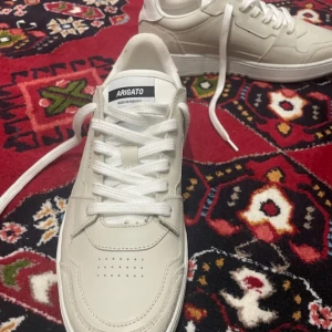 Vita sneakers från Axel Arigato - Snygga vita sneakers från Axel Arigato med stilren design. Skorna har snörning och perforerade detaljer på ovansidan. Perfekta för en casual look.