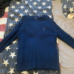Mörkblå stickad tröja från GANT - Säljer en snygg mörkblå stickad tröja från GANT. Tröjan har ett klassiskt flätmönster och rund halsringning. Perfekt för kyligare dagar. Passar både till jeans och chinos för en stilren look.