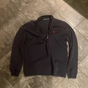 Svart tröja med dragkedja från Ralph Lauren - Snygg svart tröja från Ralph Lauren med en halv dragkedja och röd broderad logga på bröstet. Perfekt för en stilren look. Tröjan har långa ärmar och ribbade muddar.