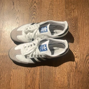 Adidas sneakers i vitt och grått - Snygga Adidas sneakers i vitt och grått med klassiska tre ränder i svart. Skorna har en blå etikett på plösen och snörning framtill. Perfekta för en avslappnad stil.