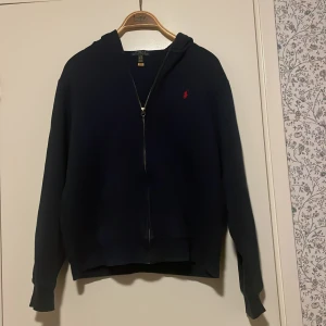 Mörkblå hoodie från Ralph Lauren - (Kan tänka mig gå ner i pris vid snabb affär) Säljer en snygg mörkblå hoodie från Ralph Lauren med dragkedja och det klassiska röda logotypbroderiet på bröstet. Perfekt för en stilren och avslappnad look. Använd men i gott skick, inga som helst defekter. (XL i barn storlek men S i herr)