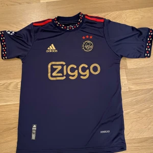 Mörkblå fotbollströja från Adidas - Säljer en mörkblå fotbollströja från Adidas med röda och guldiga detaljer. Tröjan har korta ärmar och ett mönster runt halsen och ärmsluten. Den har även en logga och sponsortryck på framsidan samt tre stjärnor ovanför klubbmärket.
