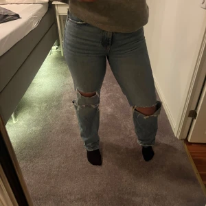 Blå jeans med slitningar - Säljer ett par blå jeans med coola slitningar på knäna. De har en hög midja och en straight passform som ger en avslappnad look. 