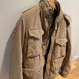 Ralph Lauren Field Jacket  - Säljer en beige field jacket från Ralph Lauren med mjukt foder inuti. Jackan har en amerikansk flagga på ärmen och axelklaffar. Perfekt för kyligare dagar med sin varma insida.
