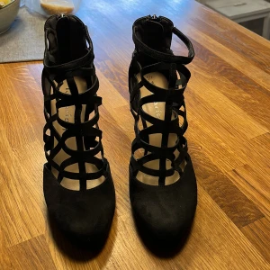Svarta klackskor från Tamaris - Snygga svarta klackskor från Tamaris med ett unikt gallerliknande mönster och dragkedja baktill. Skorna har en spetsig tå och är tillverkade i mocka, vilket ger en elegant look. Perfekta för en kväll ute eller en speciell tillställning.