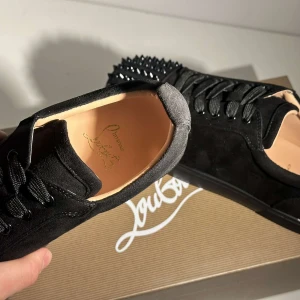  Christian Louboutin Skor - Snygga svarta sneakers i mocka från Christian Louboutin med ikoniska röda sulor. Skorna har svarta nitar på tån och snörning framtill. Perfekta för att ge en edgy touch till din stil.