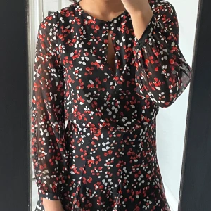 Svart klänning - Snygg svart klänning med mönster i rött och vitt. Den har långa ärmar och en liten öppning vid halsen. 🌼