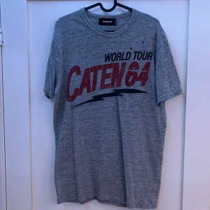 Dsquared2 T shirt - Säljer en grå t-shirt från Dsquared2 med trycket 'World Tour Caten 64' i rött och svart. T-shirten har en avslappnad passform och är kortärmad. Perfekt för en cool och avslappnad stil.