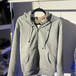Grå hoodie från Burberry - Säljer en stilren grå hoodie från Burberry med dragkedja och broderad logga på bröstet. Insidan har det klassiska Burberry-mönstret. Perfekt för en avslappnad och trendig look. Står storlek M passar som S.