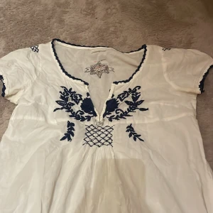 Vit blus från Odd Molly - Fin vit blus från Odd Molly med blå broderade detaljer och v-ringning. Blusen har korta ärmar med blå kant och ett fint mönster framtill. 