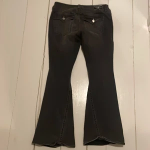 Svarta bootcut jeans från True Religion - Snygga svarta bootcut jeans från True Religion, modell Joey Low Rise Flare. De har en låg midja och är tillverkade i ett slitstarkt material med dekorativa knappar på bakfickorna. Jag ÄLSKAR dem men de är tyvärr förstora.