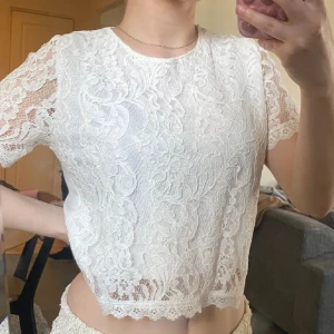 Vit spetsblus - Säljer en söt vit spetsblus med korta ärmar och ett vackert blommönster. Blusen är croppad och har en fin kantdetalj längst ner. Perfekt för en somrig look!