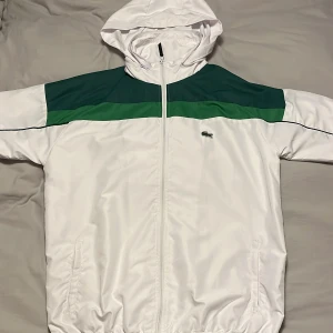 Lacoste tracksuit windbreaker - Snygg träningsdräkt från Lacoste i vitt och grönt. Jackan har en dragkedja och huva, medan byxorna har elastisk midjaband,knappt använd.