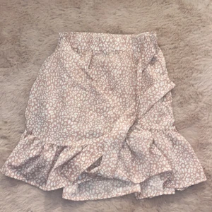 Beige/rosa leopardmönstrad omlottkjol med knytning - Snygg beige/rosa omlottkjol med leopardmönster och volangdetaljer. Perfekt för en trendig look. Kjolen har en kort längd och är gjord i ett lätt material.