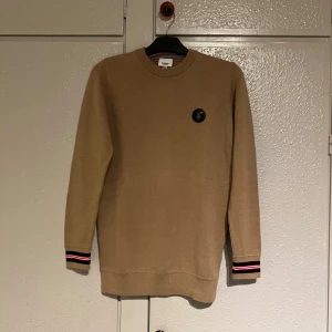 Beige tröja från Burberry - Säljer en stilren beige tröja från Burberry med rund hals och långa ärmar. Tröjan har en diskret logotyp på bröstet och ribbade muddar med röd och svart randdetalj. Perfekt för en klassisk look.