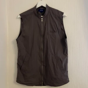 Grå vindväst från Eton - Säljer en stilren grå vindväst från Eton med dragkedja framtill. Västen har en enkel design med två fickor och en bröstficka. Perfekt för lager-på-lager-stil.