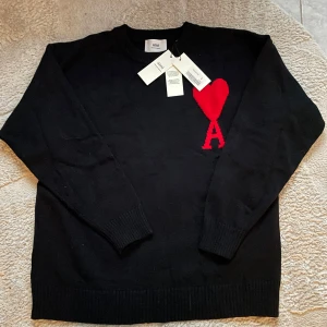 Svart tröja från Ami Alexandre Mattiussi - Säljer en stilren svart tröja från Ami Alexandre Mattiussi med ett rött hjärta och bokstaven A på bröstet. Tröjan är tillverkad av ekologisk bomull och har långa ärmar med ribbade muddar. Perfekt för en trendig look.