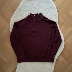 Vinröd tröja med dragkedja från Gant - Säljer en stilren vinröd tröja från Gant med halv dragkedja och hög krage. Tröjan har långa ärmar och ribbade muddar vid ärmslut och nederkant. Perfekt för kyligare dagar. Storleken är XL.