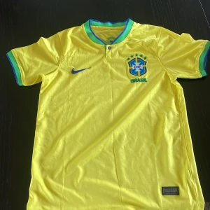 Brasilens vm tröja från 2022 - Säljer en gul Brasilien fotbollströja från Nike med korta ärmar och gröna detaljer vid krage och ärmslut. Tröjan har Dri-FIT teknologi för optimal komfort och andningsförmåga. Perfekt för fotbollsfans som vill visa sitt stöd för Brasilien! 🇧🇷