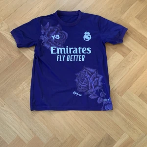 Lila Real Madrid ställ special edition  - Säljer en lila Real Madrid fotbollströja med korta ärmar. Tröjan har tryck med 'Emirates Fly Better' och 'Mbappé 9' på ryggen. Designen inkluderar blommönster och klubbens emblem på bröstet.