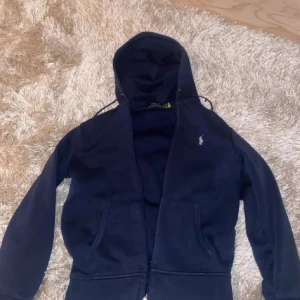 Mörkblå hoodie från Ralph Lauren - Säljer en snygg mörkblå hoodie från Ralph Lauren med dragkedja och klassisk logga på bröstet. Perfekt för en avslappnad stil. Den har en bekväm huva och långa ärmar.