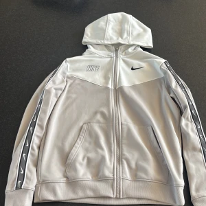 Vit hoodie från Nike - Säljer en vit hoodie från Nike med dragkedja och huva. Den har svarta detaljer och logga på bröstet samt längs ärmarna. Perfekt för en sportig look eller avslappnad stil.