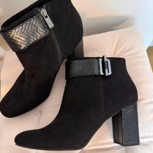 Svarta mocka boots med ormskinnsdetalj - Snygga svarta boots i mocka med en elegant ormskinnsdetalj runt ankeln och dragkedja på sidan. De har en robust klack och en stilren design som passar perfekt för höst och vinter. Används en gång. Original pris på ca 2000 kr