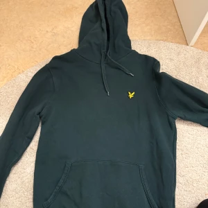 Mörkgrön hoodie från Lyle & Scott - Säljer en stilren mörkgrön hoodie från Lyle & Scott med en gul logotyp på bröstet. Tröjan har en klassisk känguruficka och justerbar huva med dragsko. Perfekt för en avslappnad look.