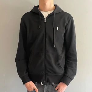 Svart hoodie från Polo Ralph Lauren - Säljer en stilren svart hoodie från Polo Ralph Lauren med dragkedja och det klassiska logotypbroderiet på bröstet. Tröjan har långa ärmar och en praktisk huva med snörning. Perfekt för en avslappnad look.  Skick 10/10 helt oanvänd.