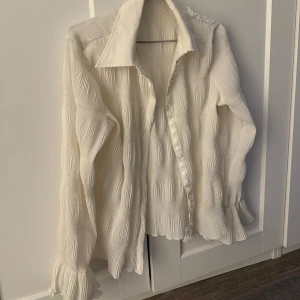 Vit blus med volangdetaljer - Säljer en elegant vit blus med volangdetaljer vid ärmslut och nederkant. Blusen har långa ärmar och knappar framtill. Perfekt för en stilren look.