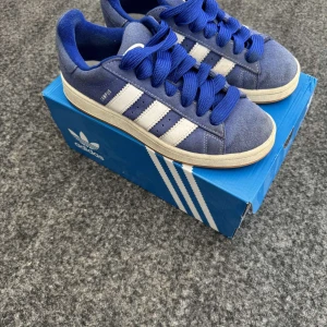 Blå Adidas Campus sneakers - Snygga blå Adidas Campus sneakers med vita ränder och snörning. Skorna har en klassisk design med gummisula och är perfekta för en avslappnad stil. De är tillverkade i mocka och har en bekväm passform. Storlek 40. Hör av dig vid frågor eller funderingar✅.