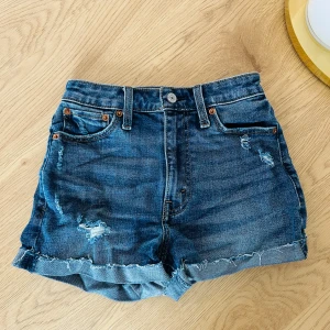 Jeansshorts från Abercrombie & Fitch - Snygga högmidjade jeansshorts från Abercrombie & Fitch i en klassisk blå denim. Shortsen har slitna detaljer och uppvikta kanter för en avslappnad look. Perfekta för varma sommardagar!