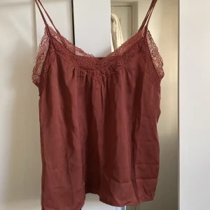 Brun/rött linne från Vero Moda - Säljer ett snyggt brunt linne från Vero Moda med spetsdetaljer vid halsringningen och tunna axelband.
