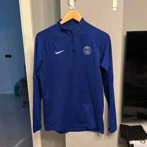 Blå långärmad träningströja från Nike - Säljer en blå långärmad träningströja från Nike med Paris Saint-Germain-logga på bröstet. Tröjan har en halv dragkedja framtill och är perfekt för fotbollsträning. Den är tillverkad i ett syntetmaterial som andas.