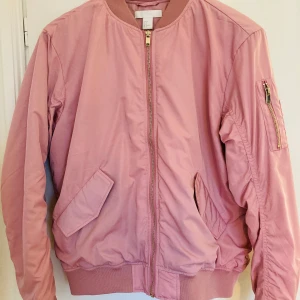 Rosa bomberjacka - Säljer en snygg rosa bomberjacka från H&M. Jackan har en dragkedja framtill och en extra ficka med dragkedja på ärmen. Tillverkad i 100% polyester, perfekt för vår och höst. Den har en klassisk bomberstil med ribbade muddar vid ärmslut och nederkant.