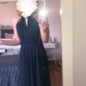 Mörkblå långklänning - Elegant mörkblå långklänning med halterneck och markerad midja. Perfekt för festliga tillfällen. Klänningen har en stilren design med ett lätt fall.