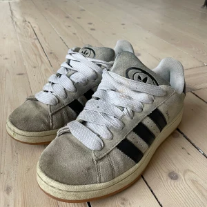 Adidas Campus sneakers i beige mocka - Säljer ett par klassiska Adidas Campus sneakers i beige mocka med svarta ränder. Skorna har en vit sula och vita snören. Perfekta för en avslappnad stil.