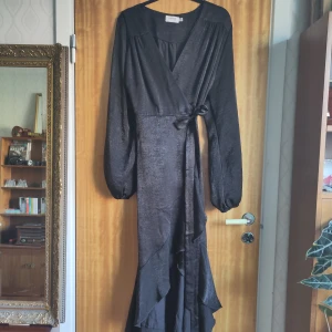 Svart omlottklänning från Flounce London - Elegant svart omlottklänning från Flounce London med långa ärmar och volangdetaljer. Klänningen har en v-ringad design och knytning i midjan för en smickrande passform. Perfekt för en stilfull look. Endast använd en gång.