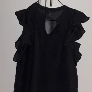 Svart blus med volangdetaljer från Vero Moda - Elegant svart blus med volangdetaljer längs ärmarna. Blusen har en v-ringad design och är ärmlös, vilket ger en stilren och modern look. Perfekt för att lyfta vilken outfit som helst.