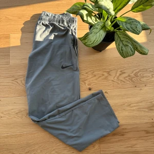 Grå träningsbyxor från Nike - Säljer ett par grå träningsbyxor från Nike med elastisk midja och Dri-FIT-teknologi för optimal komfort. Byxorna har en diskret Nike-logga på benet och är perfekta för träning eller avslappning.