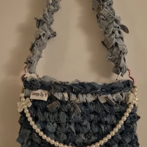 Unik virkad handväska från Loops By V - Säljer en unik virkad handväska från Loops By V. Väskan är tillverkad av återvunnet jeansmaterial med en cool, ruffig look. Den har ett pärlband som detalj och en robust axelrem. Perfekt för dig som vill vara i blickfånget.